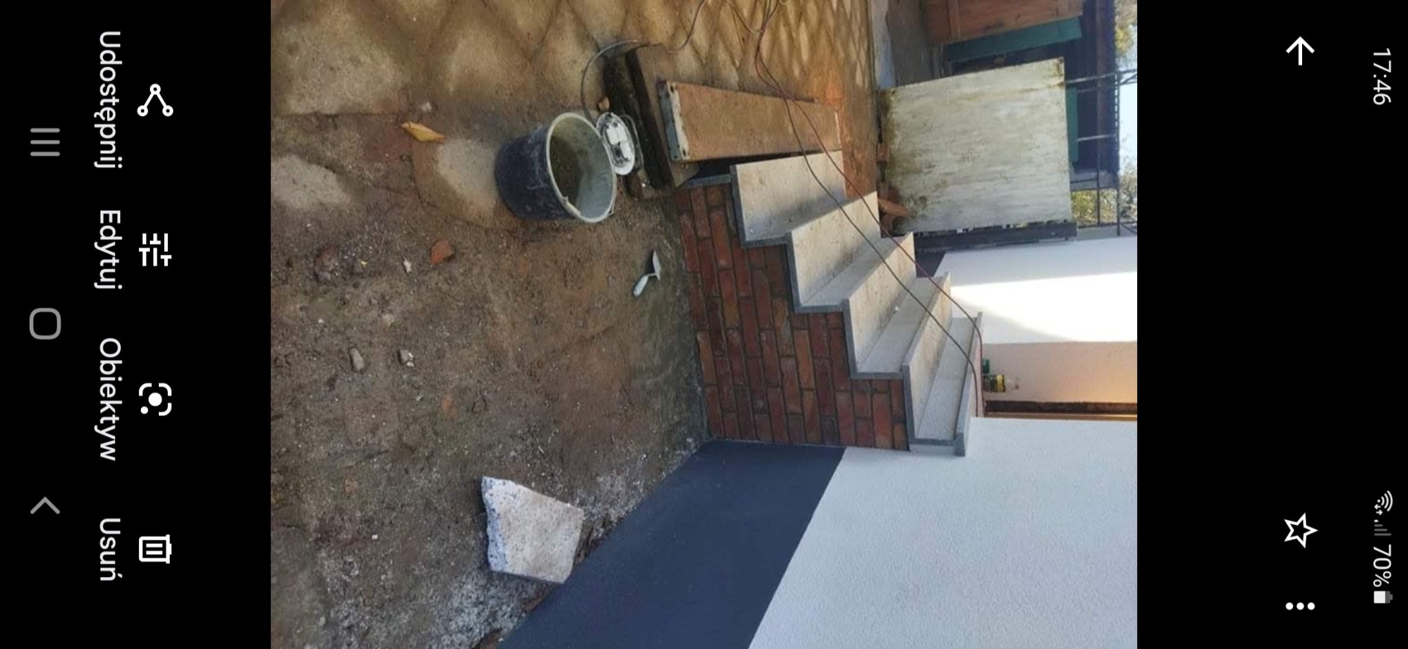 Murowane schody zewnętrzne z betonowymi stopniami w trakcie budowy, widoczny kubeł z zaprawą i kielnia na ziemi.