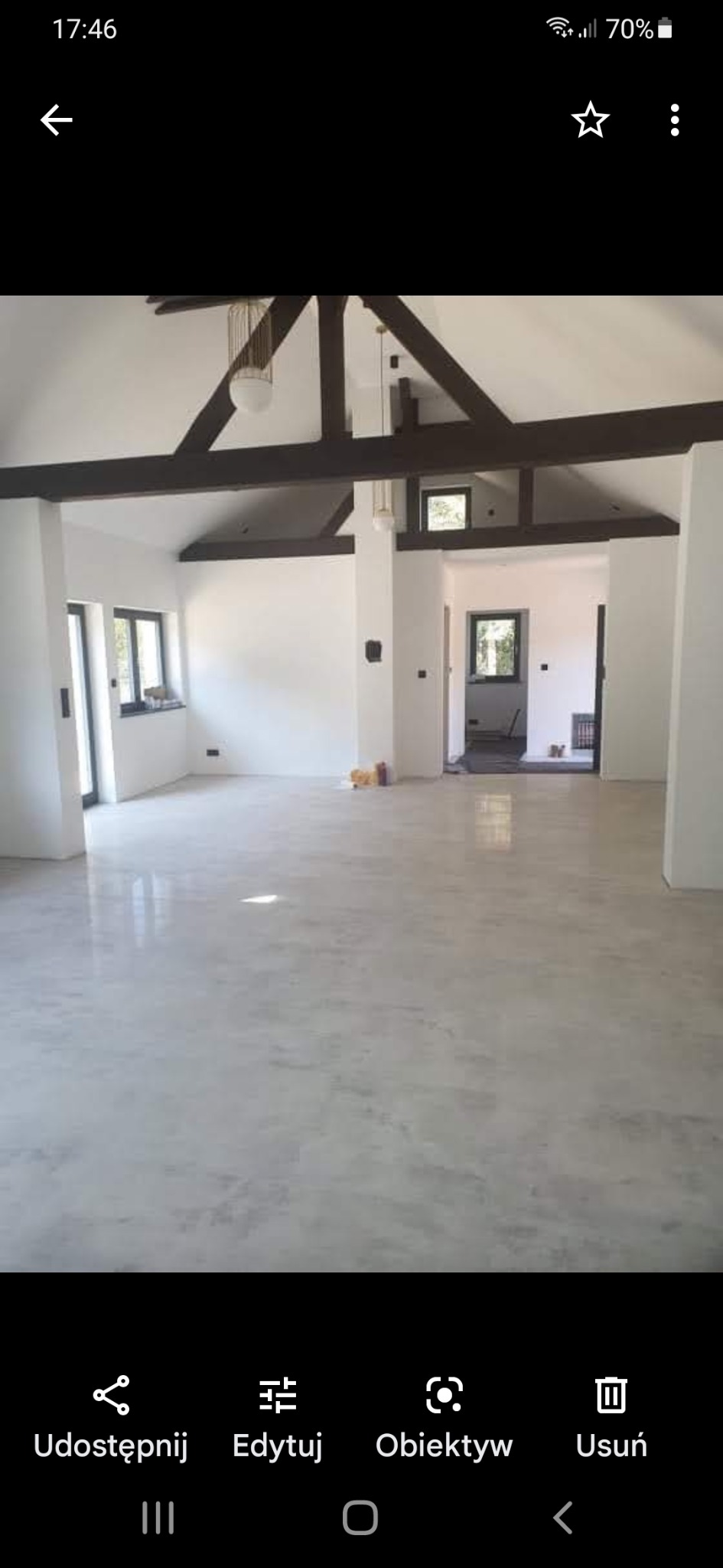 Surowe wnętrze domu w trakcie wykańczania, z widocznymi elementami konstrukcyjnymi dachu, świeżo wylana posadzka, białe ściany i czarne ramy okienne.