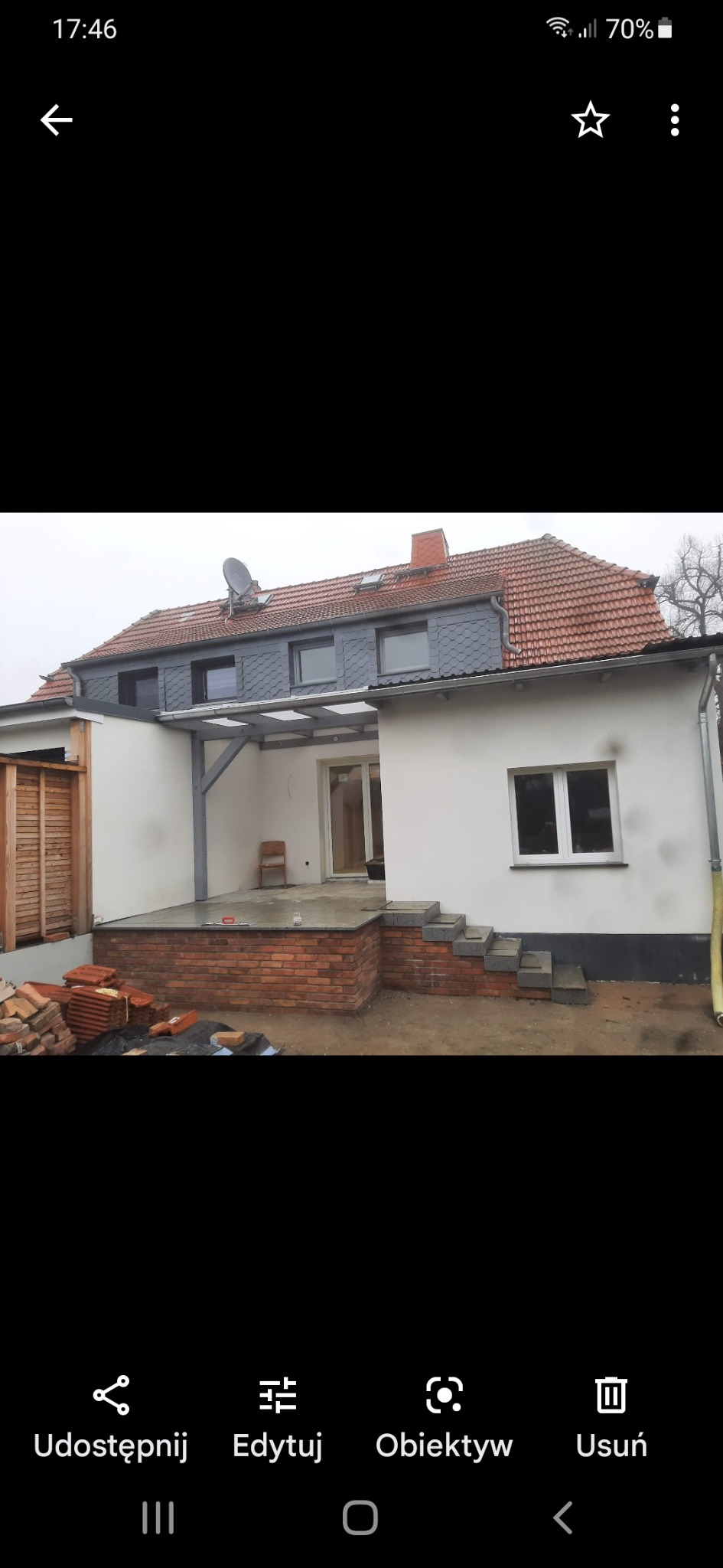 Widok z zewnątrz na dobudowany do domu taras z ceglaną ścianką i schodami z szarego kamienia, częściowo zadaszony metalową konstrukcją, z widocznymi dachówkami na ziemi podczas prac wykończeniowych.