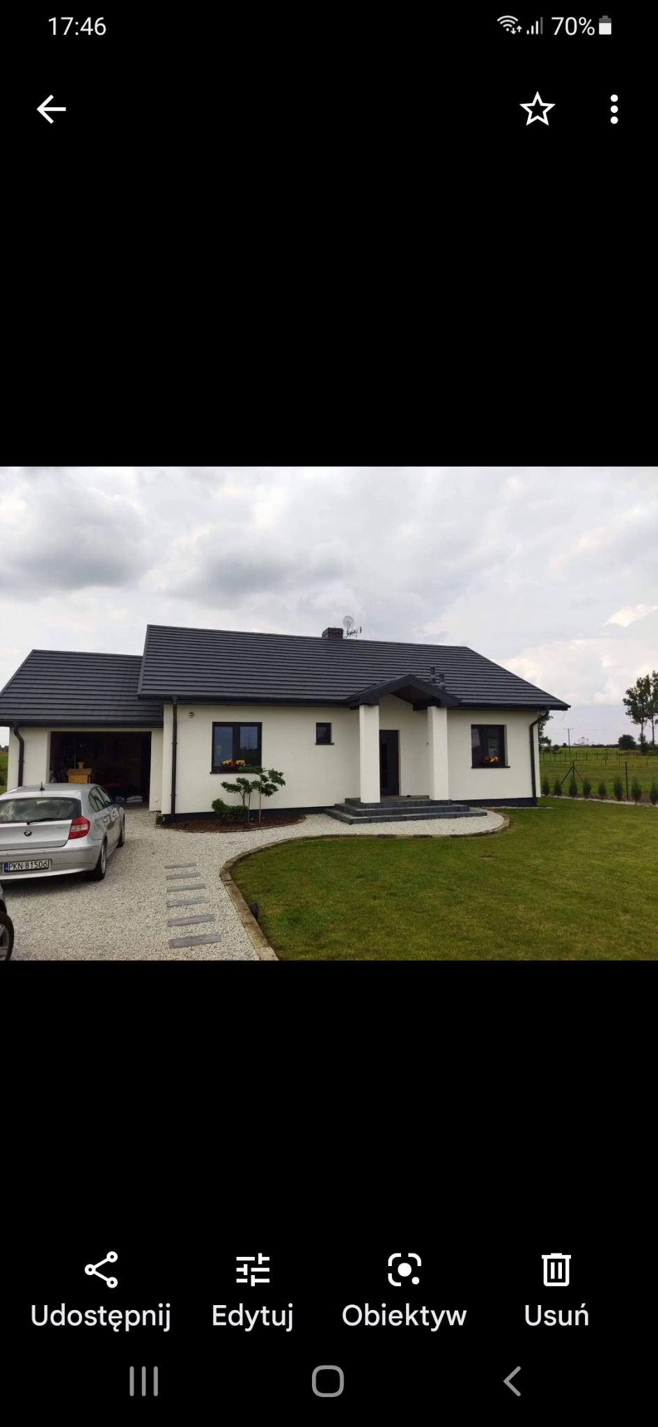 Jednorodzinny dom z jasną elewacją, ciemną dachówką i zagospodarowanym terenem wokół, widok z przodu na tle pochmurnego nieba.