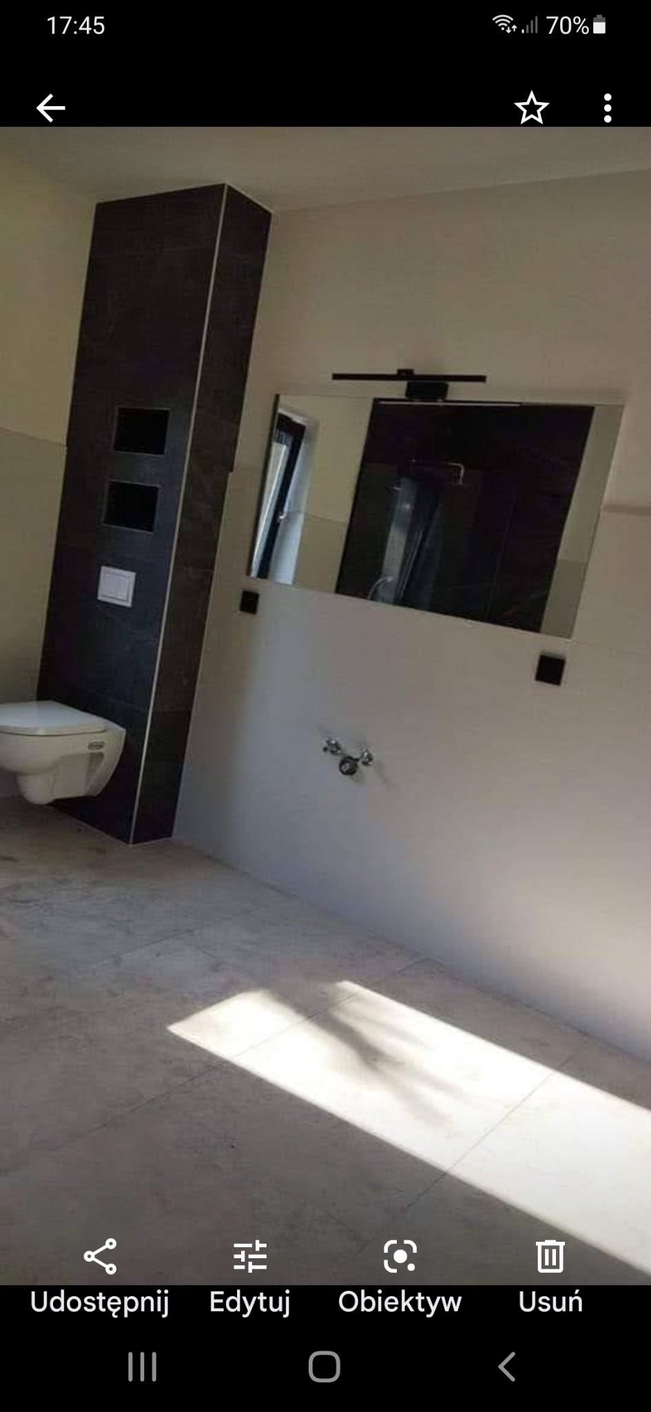 Nowoczesna łazienka z podwieszaną toaletą obudowaną ciemnymi płytkami, duże lustro z oświetleniem nad umywalką, jasna podłoga z widocznym fragmentem promieni słonecznych.