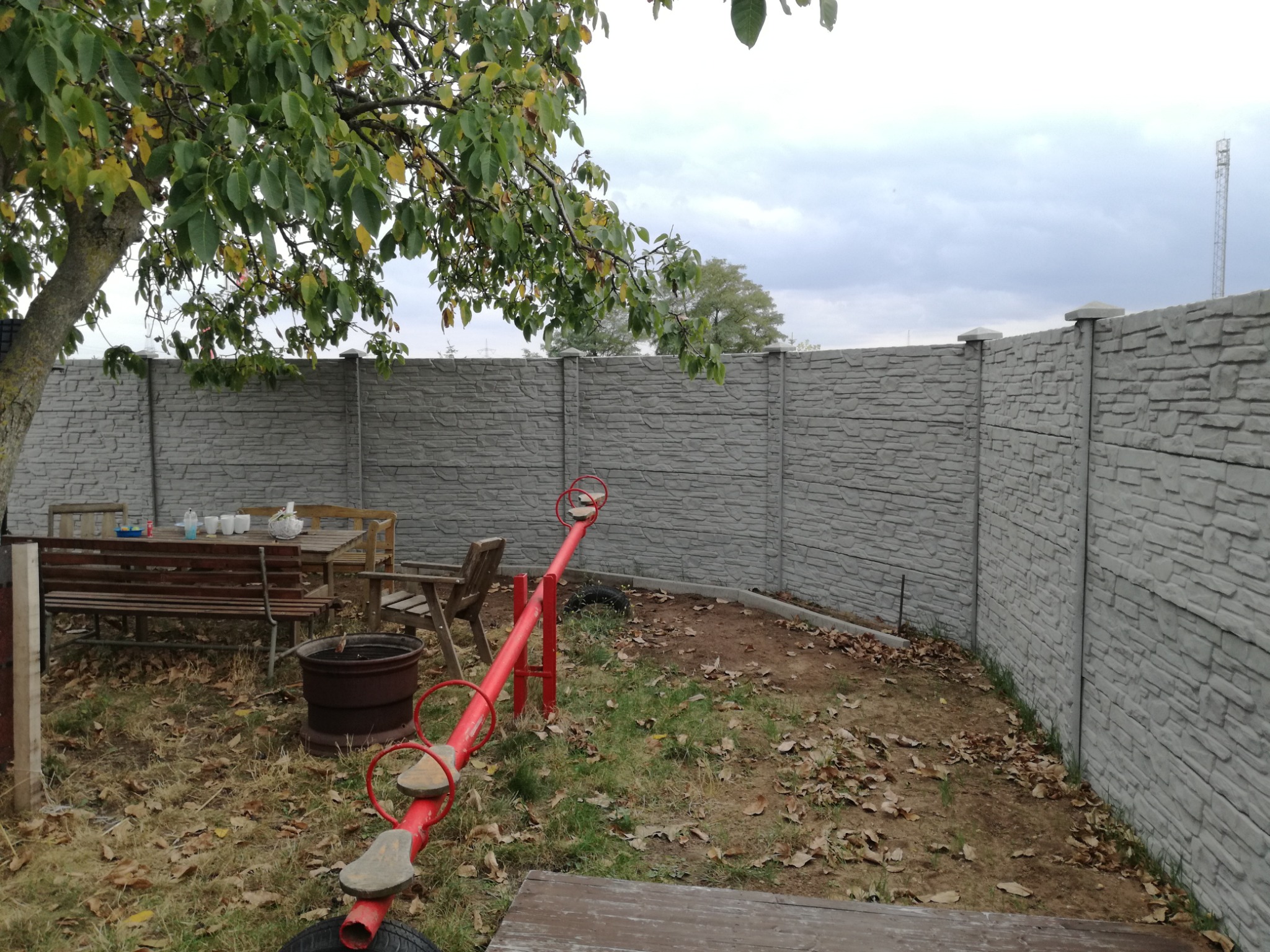 Szary betonowy płot z widoczną fakturą kamienia, za którym widać drzewo i huśtawkę w ogrodzie z opadłymi liśćmi.