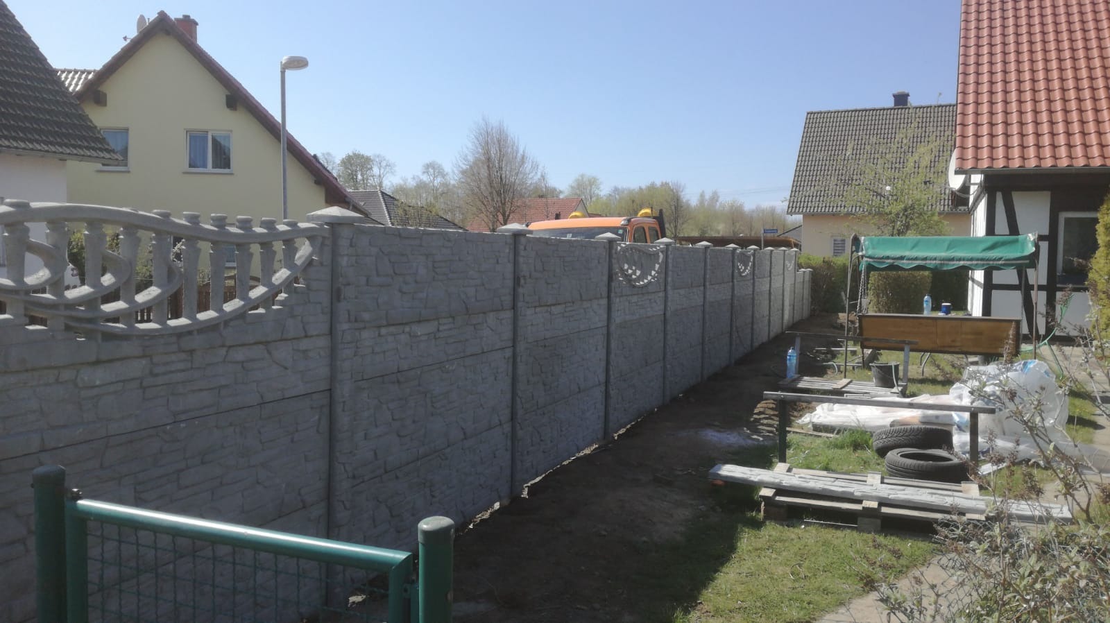 Długi, szary, betonowy płot z dekoracyjnymi elementami u góry, oddzielający posesje w Witnicy. W tle widoczny budynek mieszkalny i częściowo pomarańczowy samochód dostawczy.