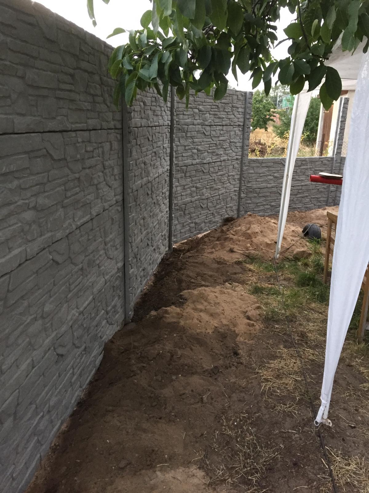 Fragment nowo postawionego, betonowego ogrodzenia panelowego imitującego kamień, z widoczną ziemią po obrysie i fragmentem białego namiotu w tle.