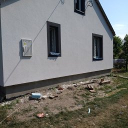 MIREMO usługi remontowo-budowlane - Świeżo otynkowana elewacja domu jednorodzinnego z trzema oknami w ciemnych obramowaniach, widoczna skrzynka elektryczna na ścianie, teren wokół domu częściowo uporządkowany, fragment anteny na dachu.
