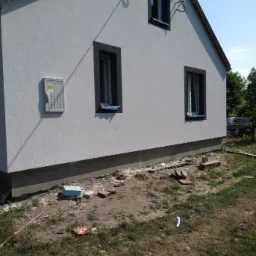 Świeżo otynkowana elewacja domu jednorodzinnego z trzema oknami w ciemnych obramowaniach, widoczna skrzynka elektryczna na ścianie, teren wokół domu częściowo uporządkowany, fragment anteny na dachu.