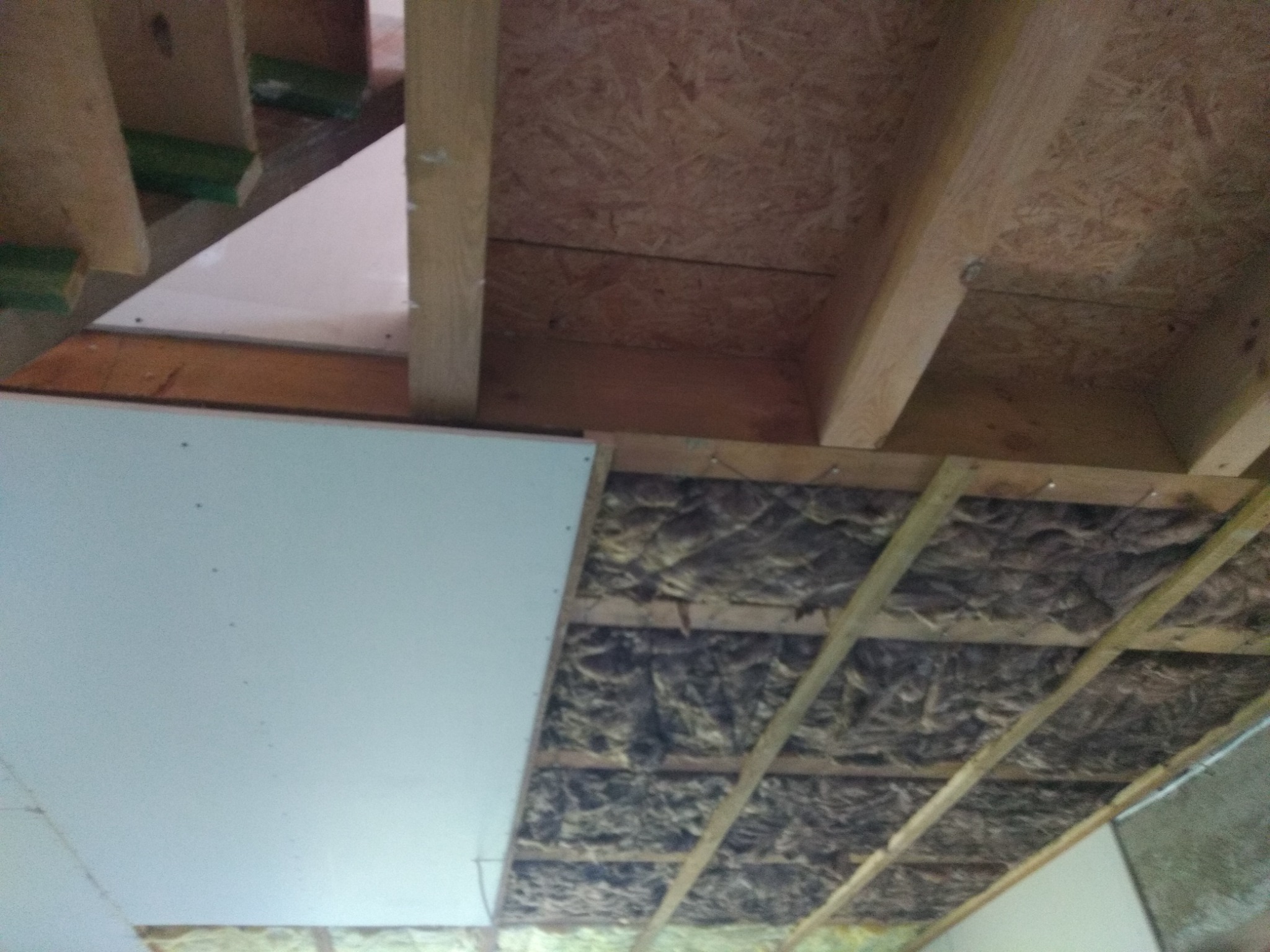 Montaż izolacji stropu wełną mineralną pomiędzy drewnianymi belkami, częściowo zakrytej płytą gipsowo-kartonową, widok od dołu.