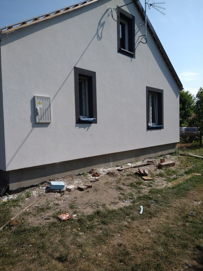 Świeżo otynkowana elewacja domu jednorodzinnego z trzema oknami w ciemnych obramowaniach, widoczna skrzynka elektryczna na ścianie, teren wokół domu częściowo uporządkowany, fragment anteny na dachu.