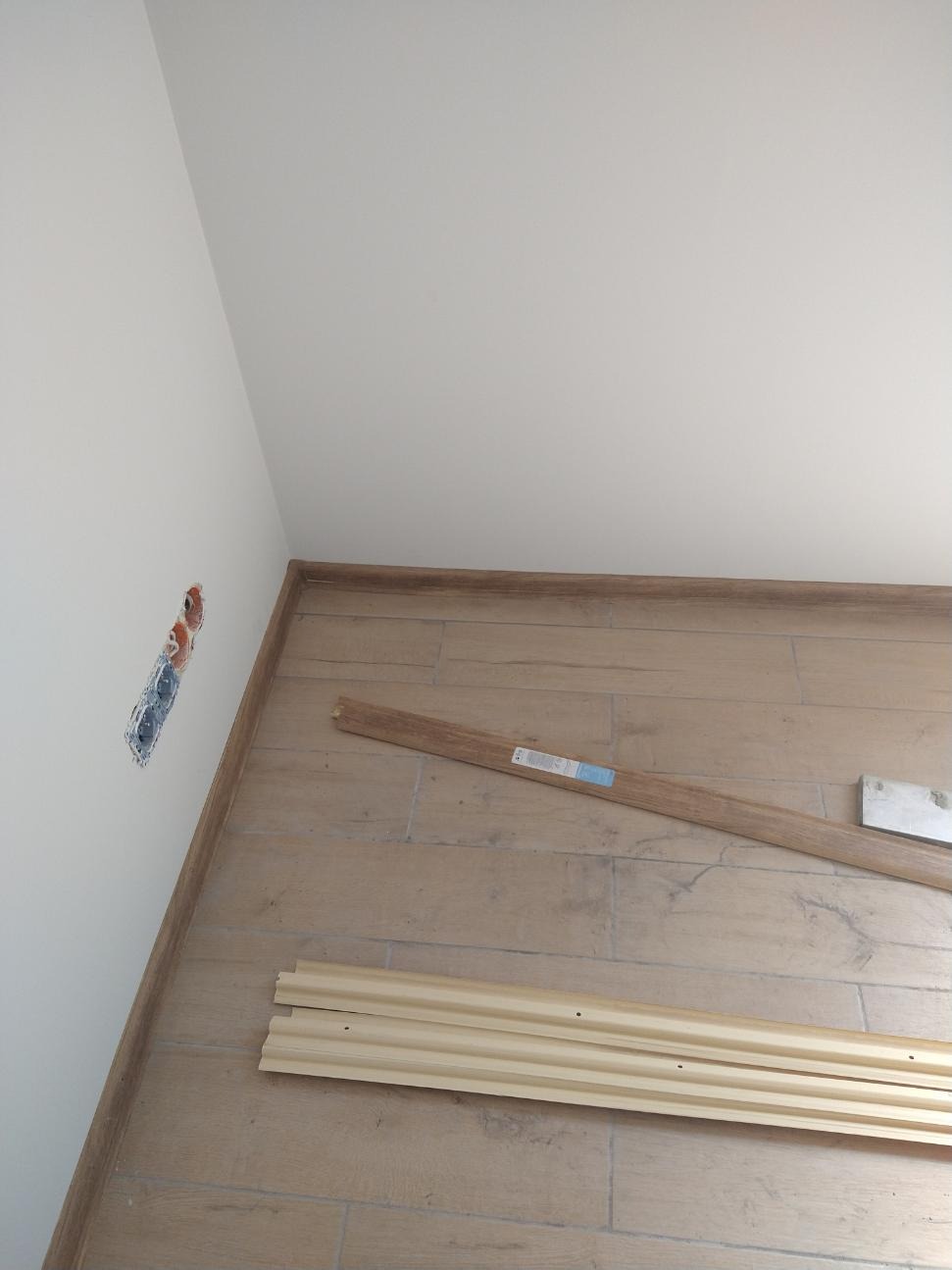 Przygotowanie do montażu drewnopodobnych listew przypodłogowych na płytkach, widoczne wycięcie w ścianie pod gniazdko elektryczne.