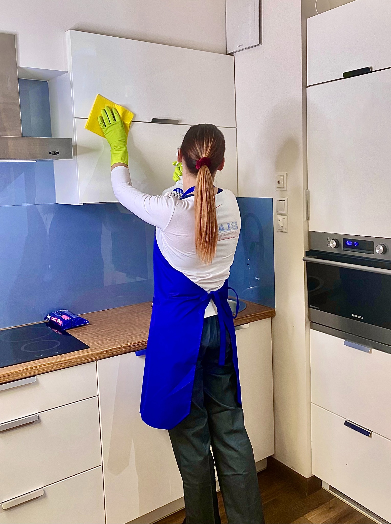 Kobieta w niebieskim fartuchu i zielonych rękawiczkach czyści żółtą ściereczką białą szafkę kuchenną; w tle widoczny okap i niebieski panel nad blatem.