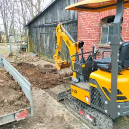 Żółta mini koparka NEX 10 z łyżką nabiera ziemię obok przyczepy wypełnionej ziemią, w tle budynek z cegły i drewniana szopa.