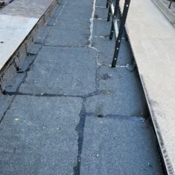 Płaski dach pokryty papą bitumiczną, widoczne łączenia i zabezpieczenia krawędzi. Metalowa barierka ochronna wzdłuż krawędzi dachu, w tle chodnik i kosze na śmieci.