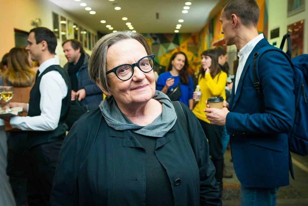 Uśmiechnięta kobieta w okularach na tle wydarzenia kulturalnego, w tle widoczni ludzie i element dekoracji ściennej.