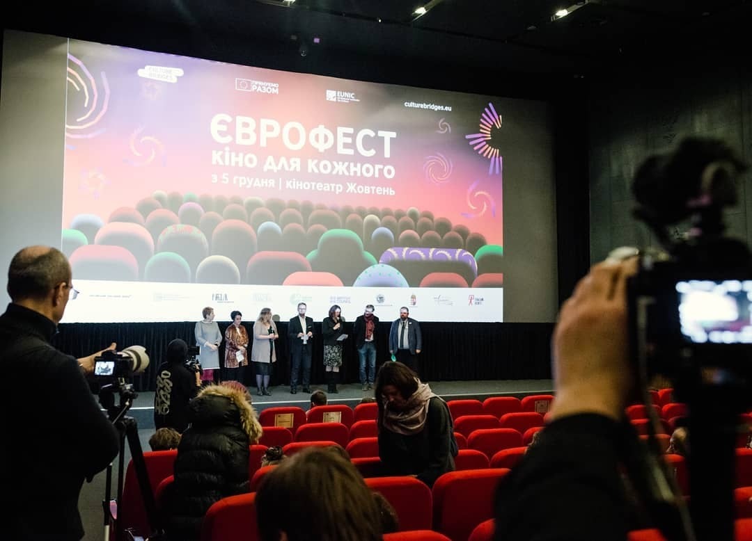 Grupa osób na scenie przed ekranem podczas festiwalu filmowego, widok z sali kinowej z osobami filmującymi wydarzenie.