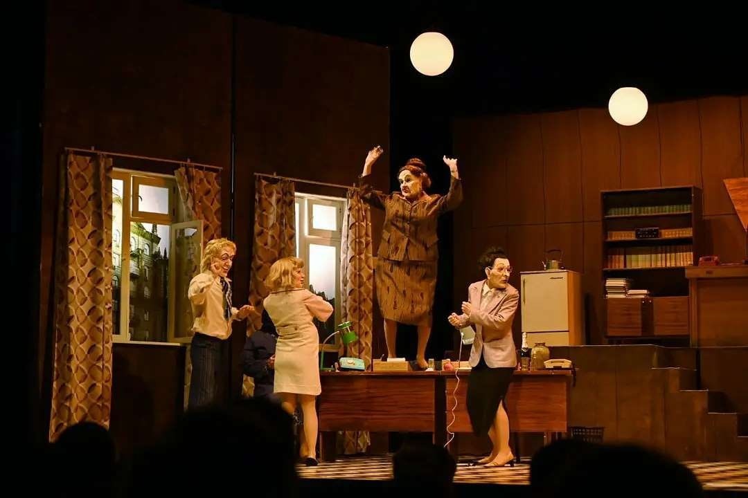Scena teatralna z aktorami w kostiumach retro, kobieta w brązowym kostiumie stoi na biurku z podniesionymi rękami, a inni aktorzy obserwują ją z boku.