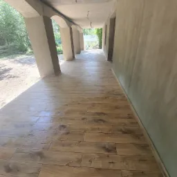 Zadaszony taras z nową drewnopodobną podłogą z płytek, surowe ściany w trakcie wykończenia, widoczne słupy podtrzymujące łukowe sklepienie.