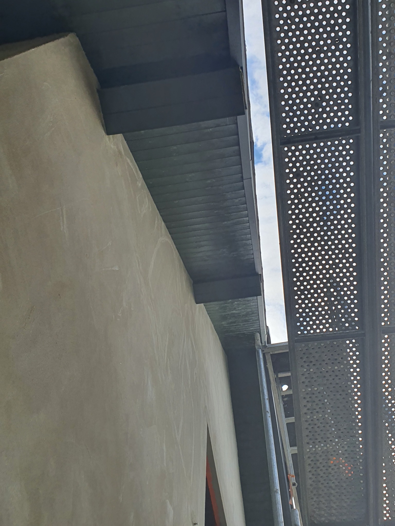 Ściana budynku z szarą elewacją w trakcie prac, widoczne rusztowanie metalowe z perforowanymi platformami, fragment podbitki dachowej w kolorze ciemnoszarym, niebo w tle.