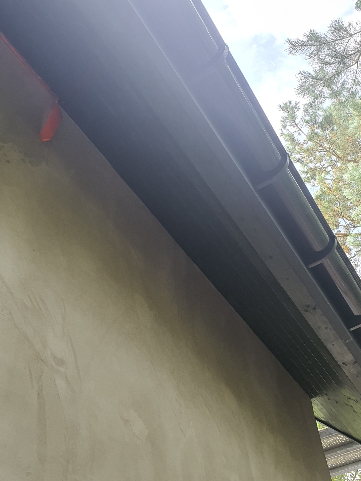 Surowa elewacja domu w trakcie prac wykończeniowych, widoczne czarne rynny i fragment rusztowania, zieleń drzew w tle.