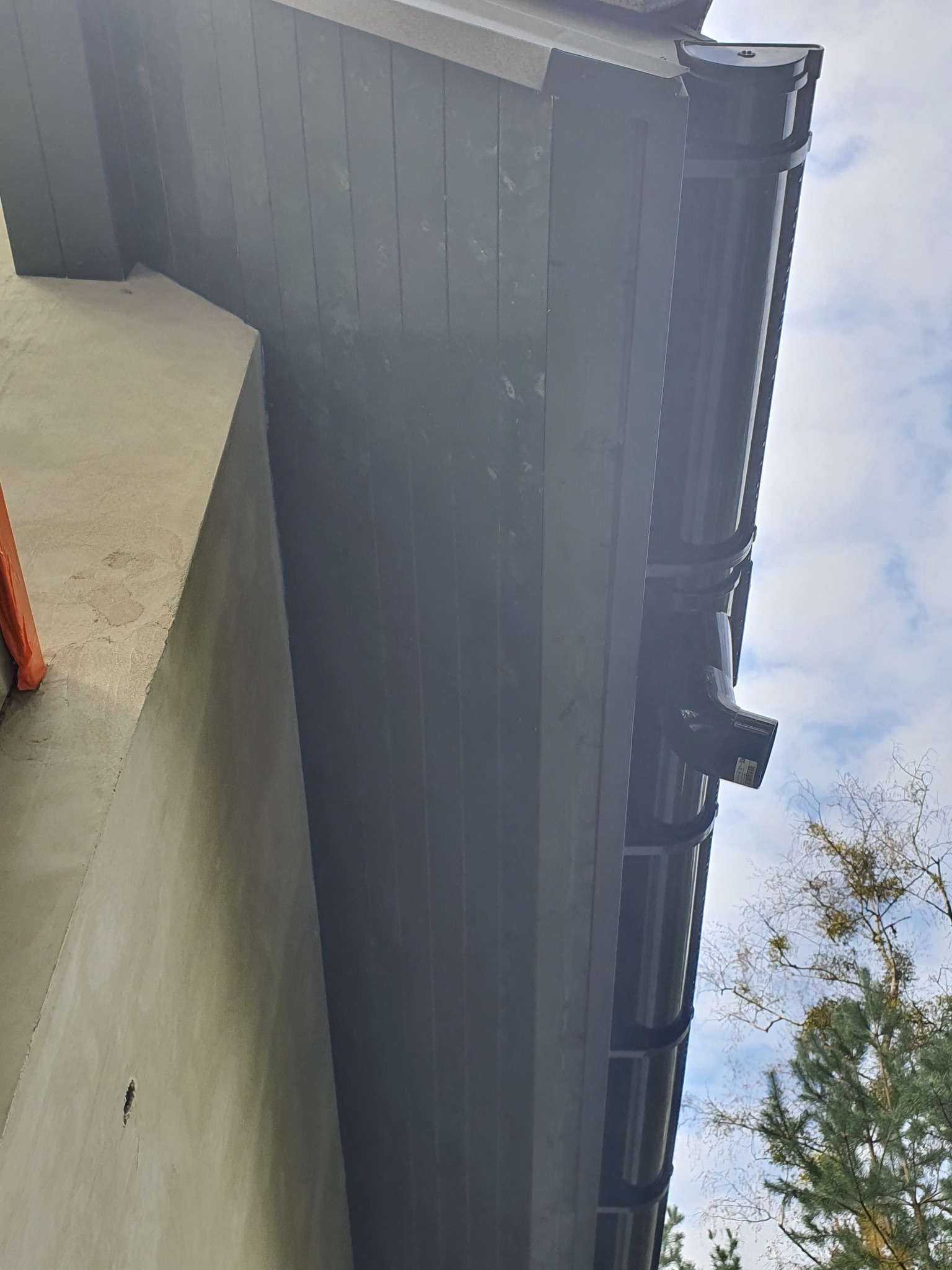 Fragment narożnika domu z zamontowaną, ciemną rynną spustową, widoczne świeżo otynkowane ściany i fragment elewacji z drewnianą boazerią w tle błękitne niebo z chmurami i korony drzew.