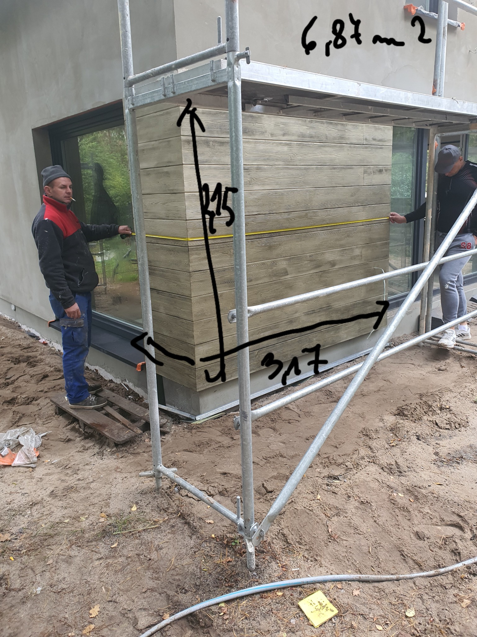 Pracownicy na rusztowaniu mierzą elewację z paneli imitujących drewno, zamontowaną na fragmencie budynku z dużym oknem.