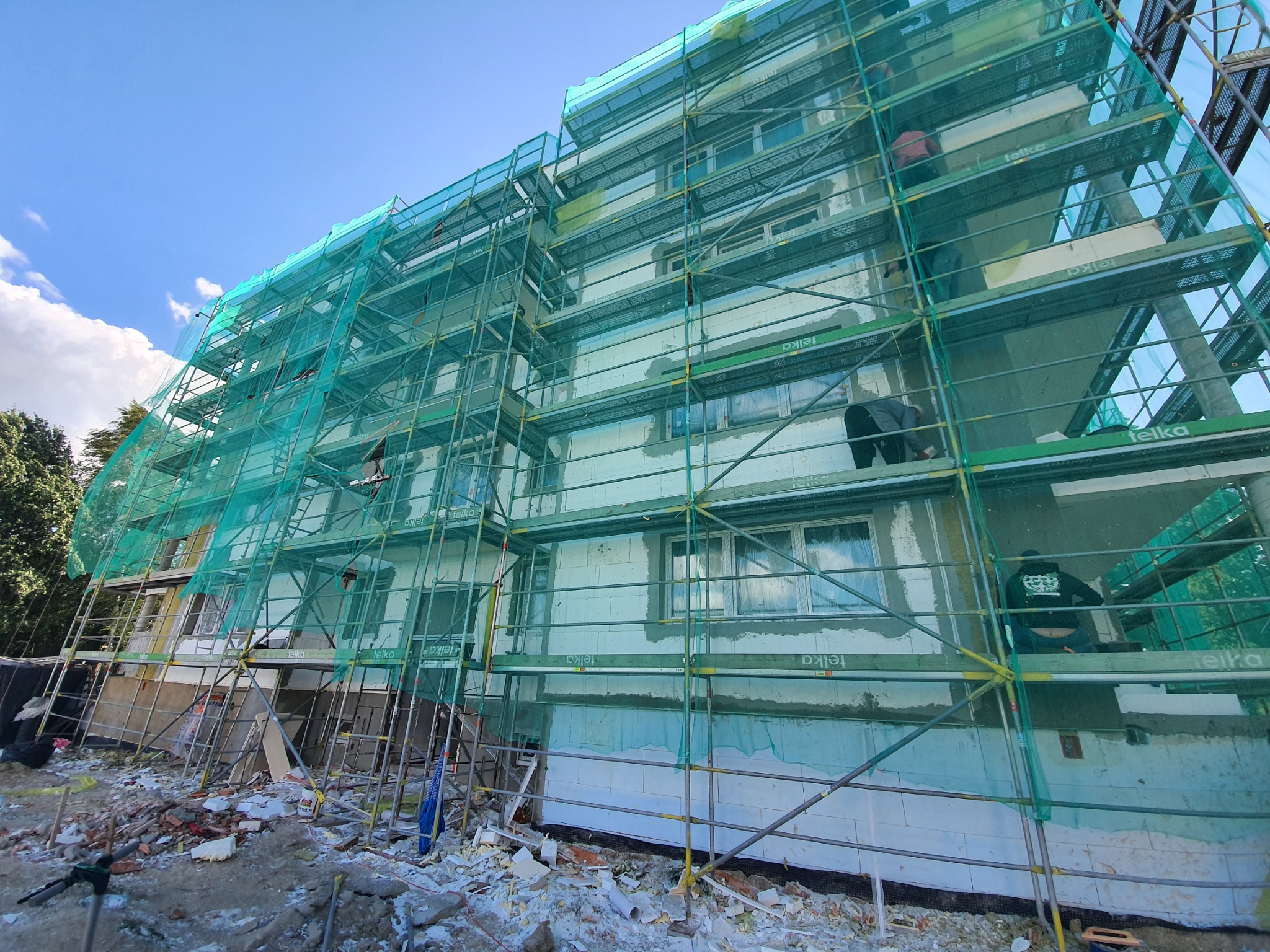 Budynek w trakcie ocieplania, widoczne rusztowania z siatką ochronną, ocieplone fragmenty elewacji, pracownicy na rusztowaniu i gruz przed budynkiem.