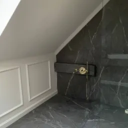 Wykończona łazienka na poddaszu ze skośnym sufitem, ściany wyłożone ciemnym marmurem z białymi żyłami, złota armatura, białe listwy przypodłogowe i dekoracyjne ramy na ścianie.