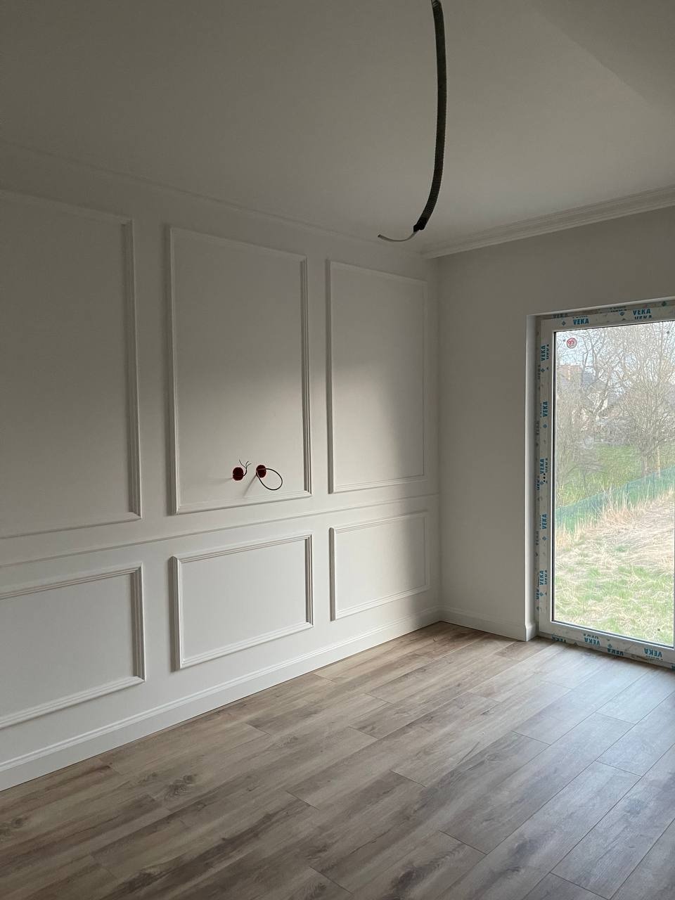 Pusty pokój po remoncie z panelami ściennymi w stylu angielskim, drewnopodobną podłogą i widoczną instalacją elektryczną na ścianie oraz przewodem wychodzącym z sufitu.