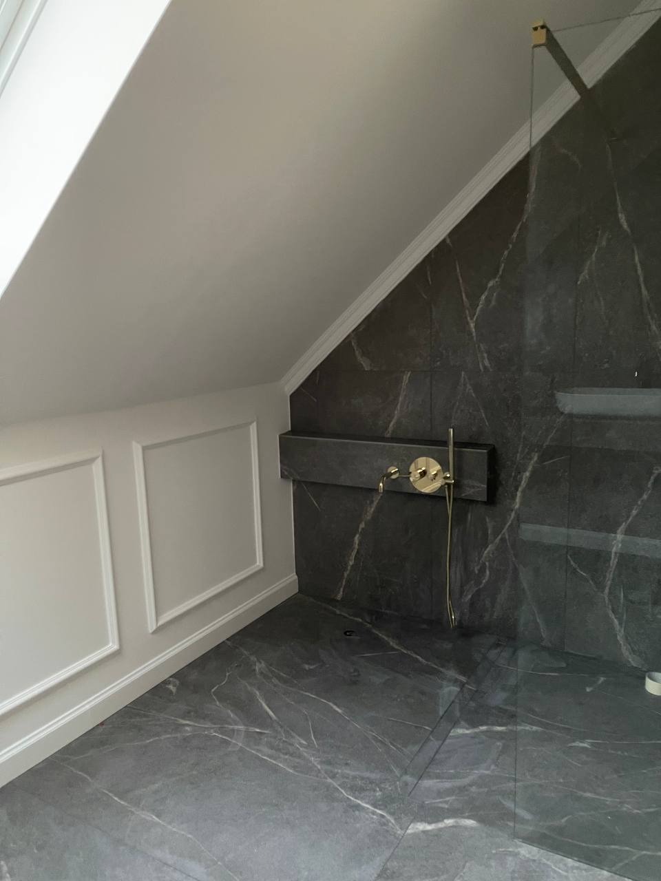 Wykończona łazienka na poddaszu ze skośnym sufitem, ściany wyłożone ciemnym marmurem z białymi żyłami, złota armatura, białe listwy przypodłogowe i dekoracyjne ramy na ścianie.