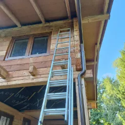 Drewniany dom w trakcie montażu rynny spustowej, widoczna drabina oparta o ścianę, brązowa rynna i fragment okna oraz zielone drzewa w tle.