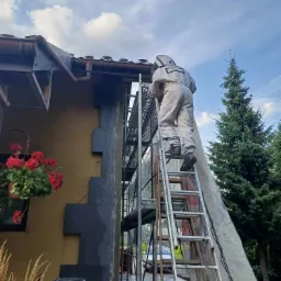 Pracownik w kombinezonie ochronnym na rusztowaniu, docieplający ścianę budynku styropianem grafitowym, widok z dołu na tle błękitnego nieba.