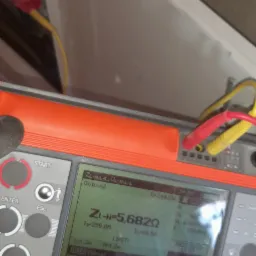 Miernik Sonel MPI-530 z podłączonymi przewodami pomiarowymi, wyświetlający wynik pomiaru impedancji pętli zwarcia ZI-N=5,682Ω, obok otwarta skrzynka elektryczna.