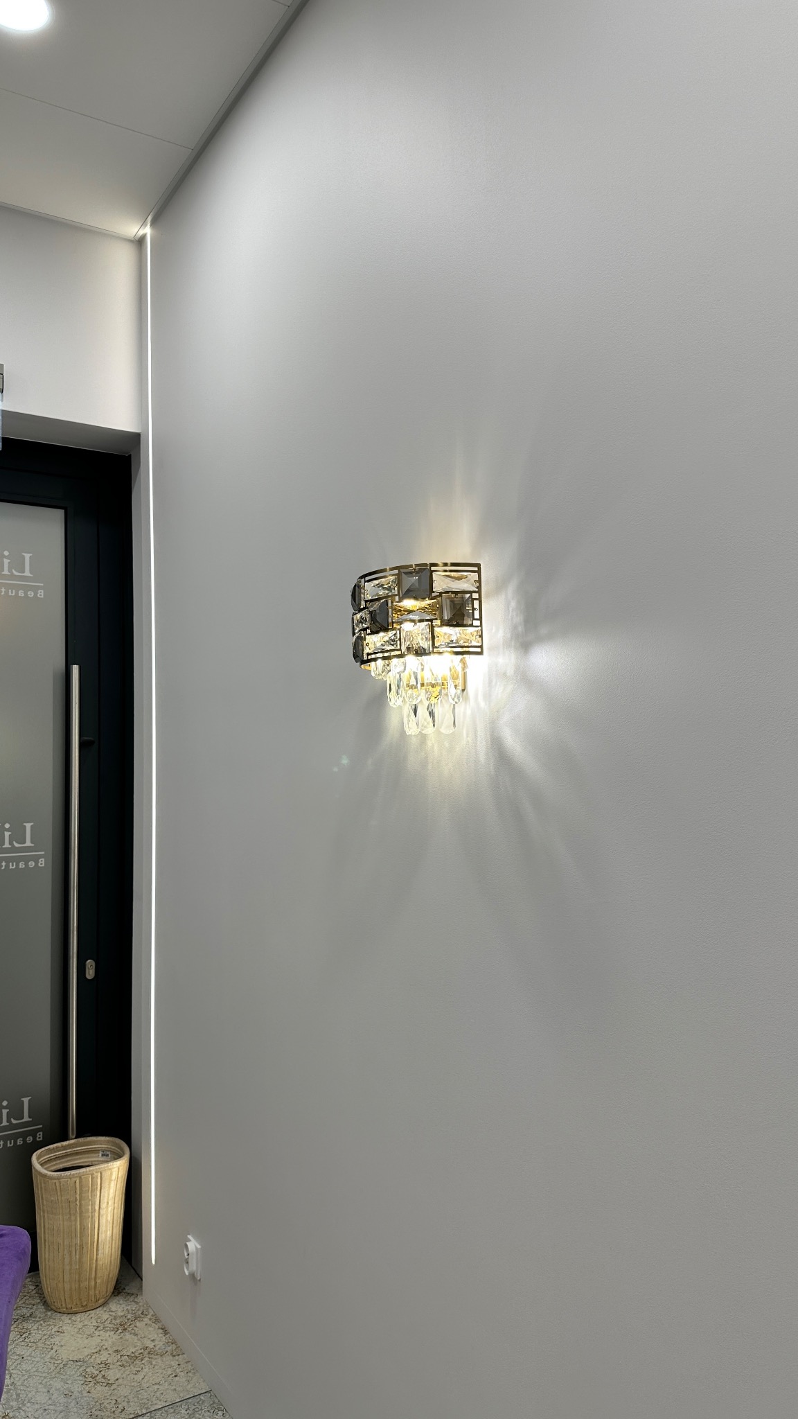 Kinkiet ścienny w stylu glamour z kryształkami, zamontowany na gładkiej, szarej ścianie w pobliżu drzwi z podświetleniem LED.