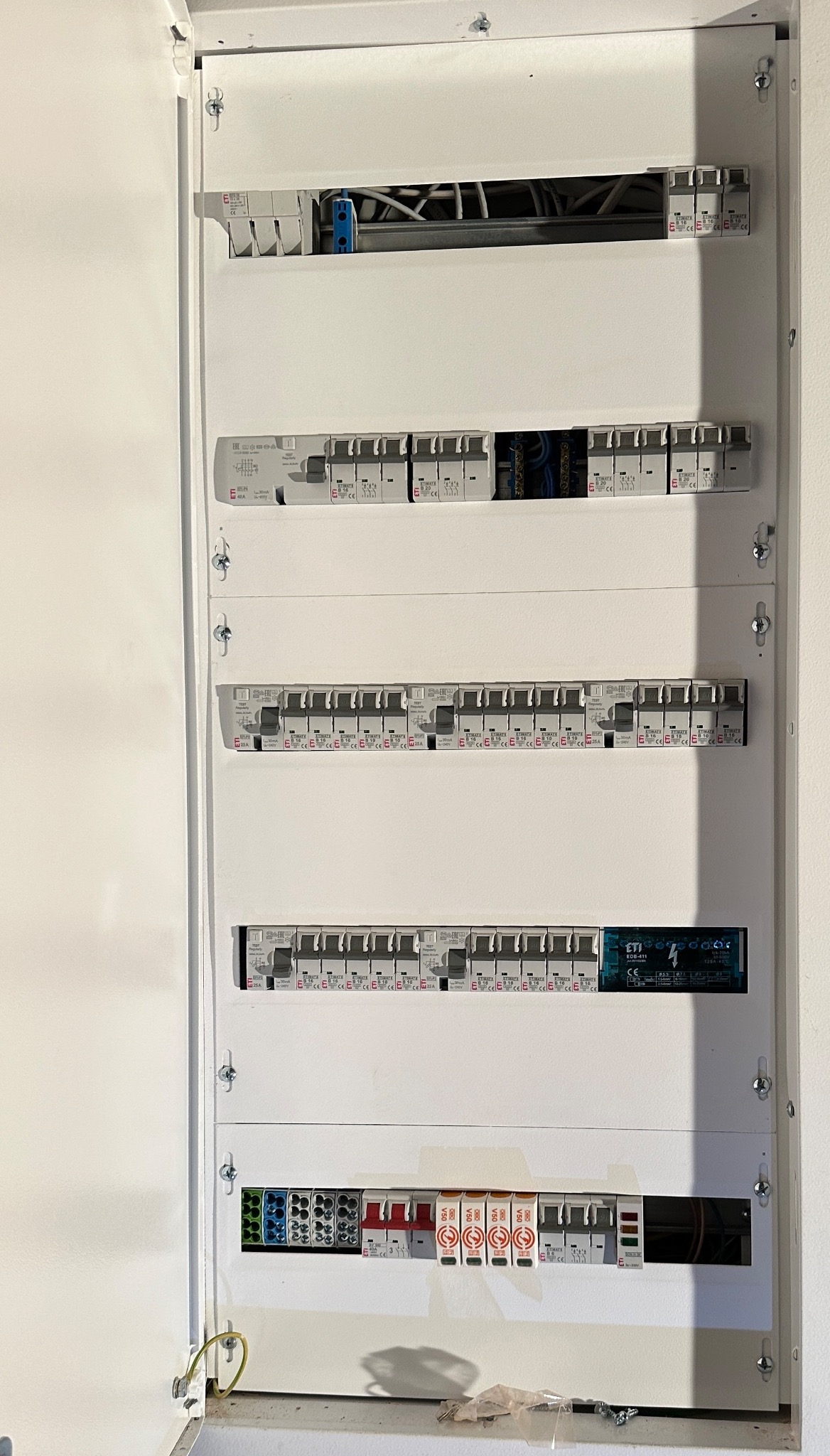 Otwarta, biała skrzynka rozdzielcza z zamontowanymi bezpiecznikami i przewodami, widoczne śruby mocujące i cień na dolnej krawędzi.