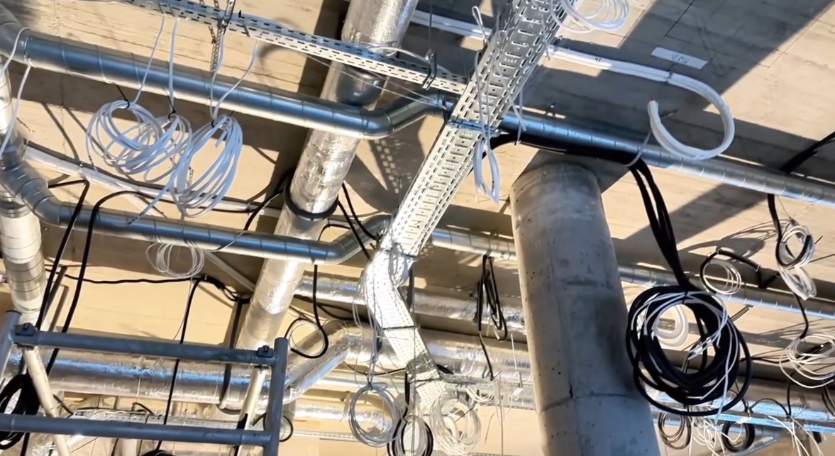 Widok z dołu na sufit z licznymi rurami, przewodami elektrycznymi i korytami kablowymi, częściowo zamontowanymi, częściowo wiszącymi, z widocznym fragmentem drabiny.