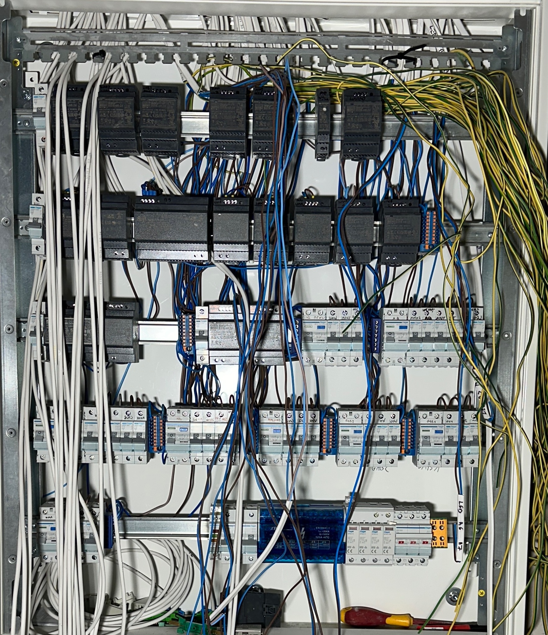 Rozdzielnica elektryczna z zainstalowanymi modułami, bezpiecznikami i przewodami w różnych kolorach, widoczny fragment śrubokręta w dolnym rogu.