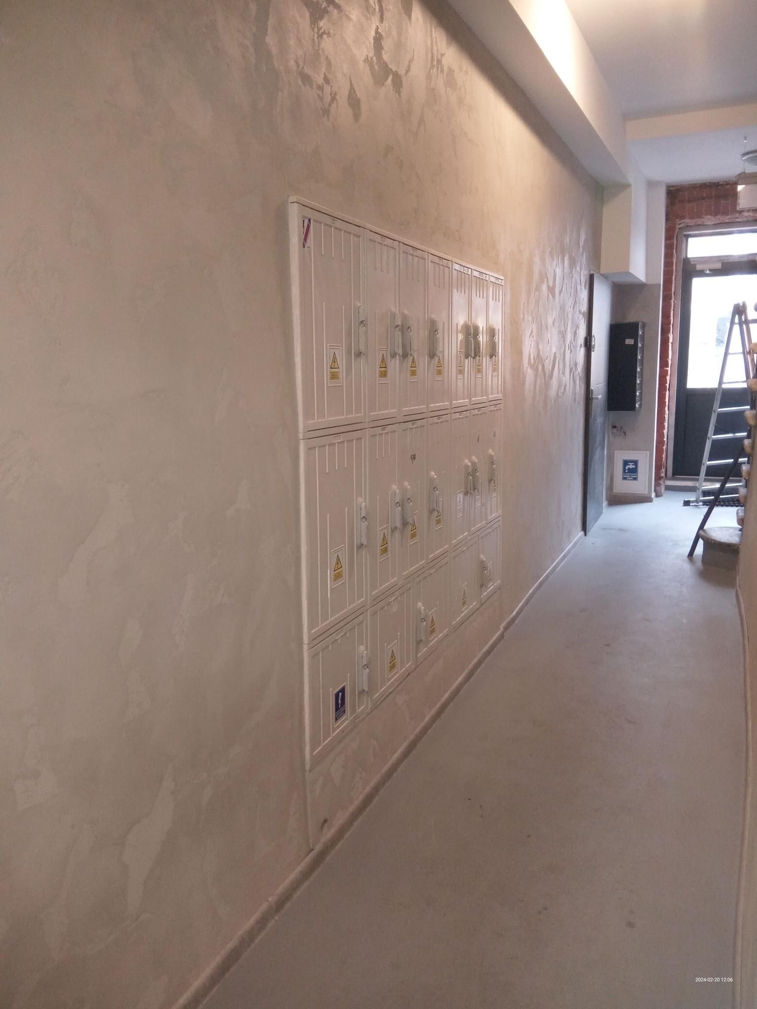 Korytarz z szafkami elektrycznymi na ścianie o dekoracyjnej fakturze, widoczna drabina w tle i drzwi wejściowe.