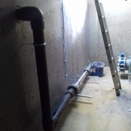 Kwinstal Krystian Wodzyński - Szara betonowa studzienka z instalacją rurową, drabina oparta o ścianę, niebieski zawór i stojąca woda na podłodze.