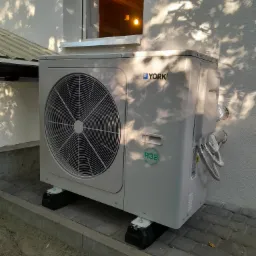 Zewnętrzna jednostka pompy ciepła marki York zamontowana na czarnych podstawach przy ścianie budynku, z widocznymi przewodami w izolacji aluminiowej i cieniem drzewa na elewacji.
