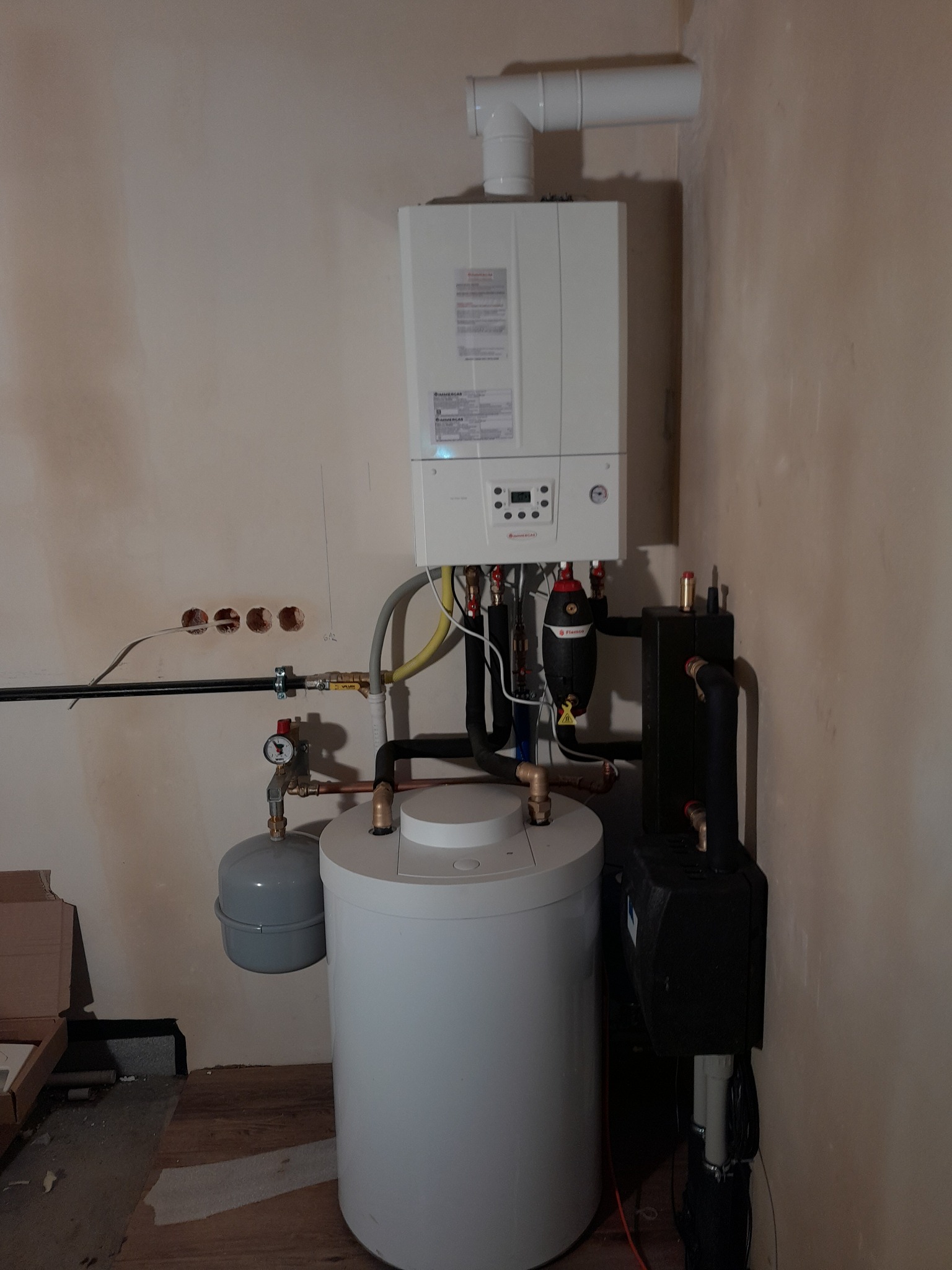 Biały piec gazowy Immergas zamontowany na ścianie w trakcie instalacji, widoczne podłączenia hydrauliczne, rury, zbiornik wyrównawczy i otwory w ścianie po instalacji.