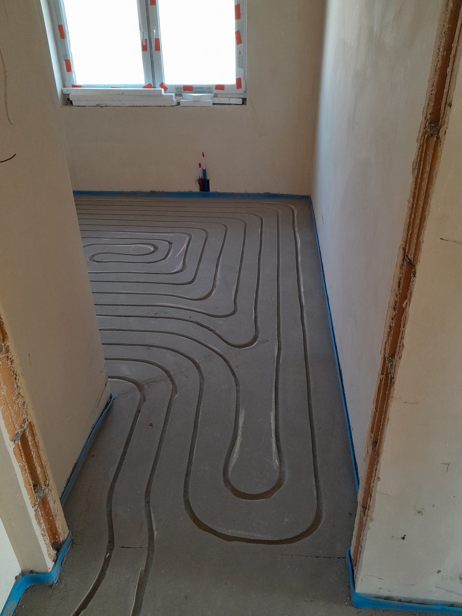 Wykonywanie instalacji ogrzewania podłogowego w nowym budynku, widoczne rury ułożone w jastrychu, zabezpieczone niebieską folią na styku ze ścianami. Okno w tle z folią ochronną.