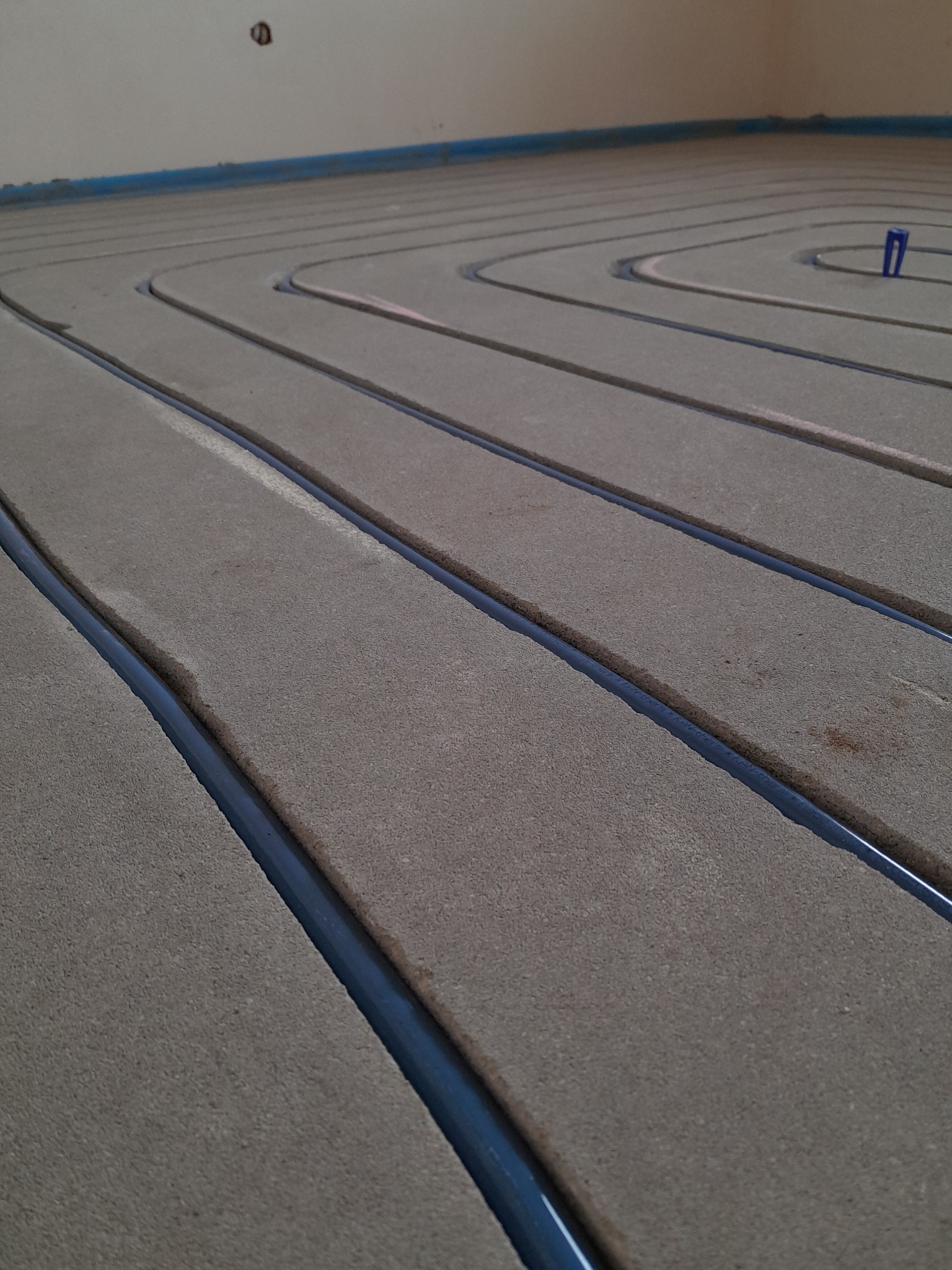 Fragment instalacji ogrzewania podłogowego w trakcie montażu, widoczne rury grzewcze ułożone w rowkach w izolacji podłogowej, z niebieskim kołkiem mocującym.