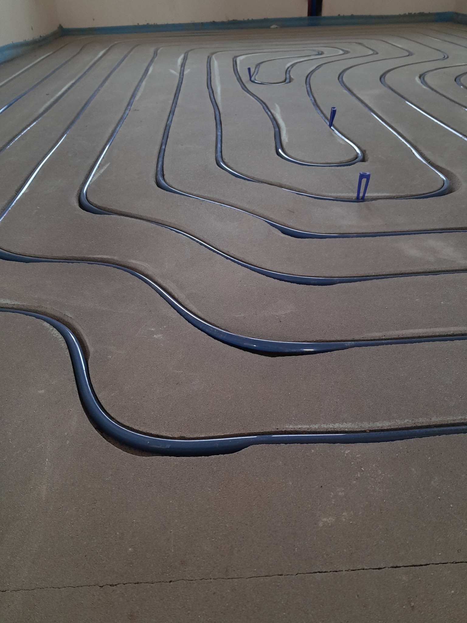 Instalacja ogrzewania podłogowego w trakcie układania w Wieliszewie, widoczne rurki w ciemnym kolorze, zamocowane do podłoża za pomocą niebieskich uchwytów.