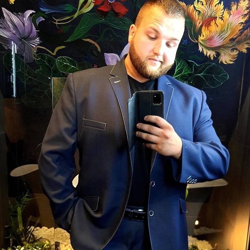 Mężczyzna w granatowym garniturze robi selfie na tle ściany z motywem kwiatowym.