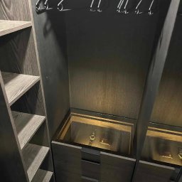 Marek Zarotyński Marq Wood Design - Wnętrze ciemnej, drewnianej garderoby z półkami i szufladami. W szufladzie podświetlonej od góry leży zegarek i portfel.