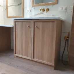 Szafka pod umywalkę z jasnego drewna z okrągłymi uchwytami na tle marmurowej ściany z złotą armaturą i lustrami w drewnianych ramach.