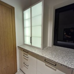 Marek Zarotyński Marq Wood Design - Jasna zabudowa kuchenna z szafkami z mlecznego szkła i blatem z szarego granitu, czarne uchwyty, widok na okno z odbiciem osoby robiącej zdjęcie.