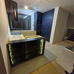 Marek Zarotyński Marq Wood Design - Nowoczesna kuchnia w trakcie montażu: czarne szafki z podświetleniem LED, białe blaty, ciemne szafy w zabudowie i okap. Widoczne elementy instalacji elektrycznej.