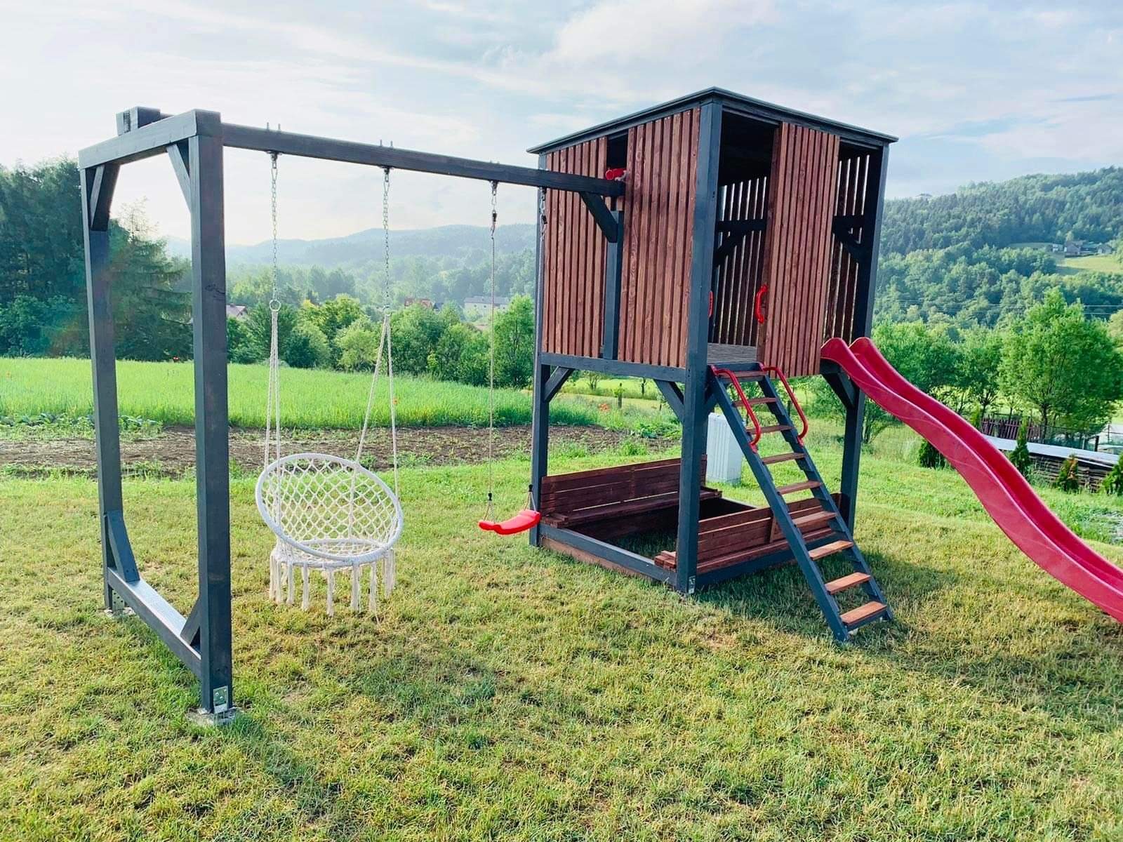 Drewniany plac zabaw z wieżą, zjeżdżalnią i huśtawką typu bocianie gniazdo na tle zielonego trawnika i zalesionych wzgórz.
