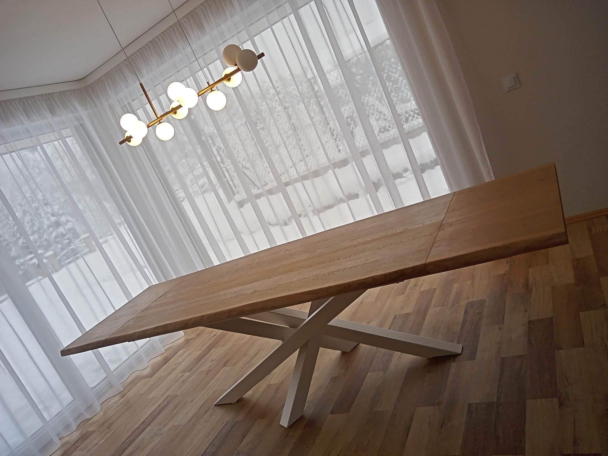 Rozkładany stół z blatem z jasnego drewna i białą, krzyżową podstawą, oświetlony nowoczesną lampą na tle okna z firanami.