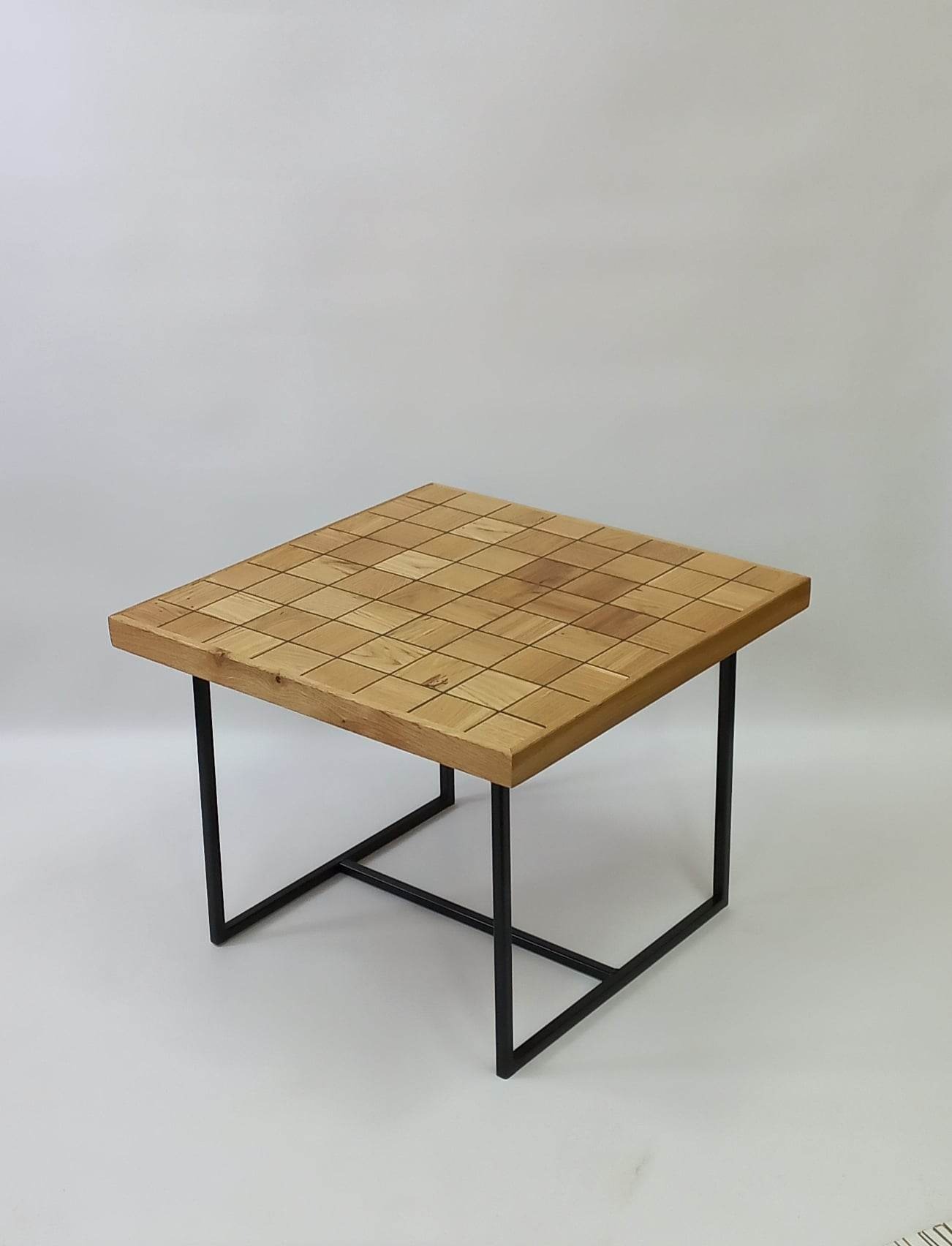 Kwadratowy stolik kawowy z blatem z jasnego drewna podzielonym na kwadraty z ciemniejszymi liniami, osadzony na czarnych, metalowych nogach w kształcie prostopadłościanu, na jasnym tle.